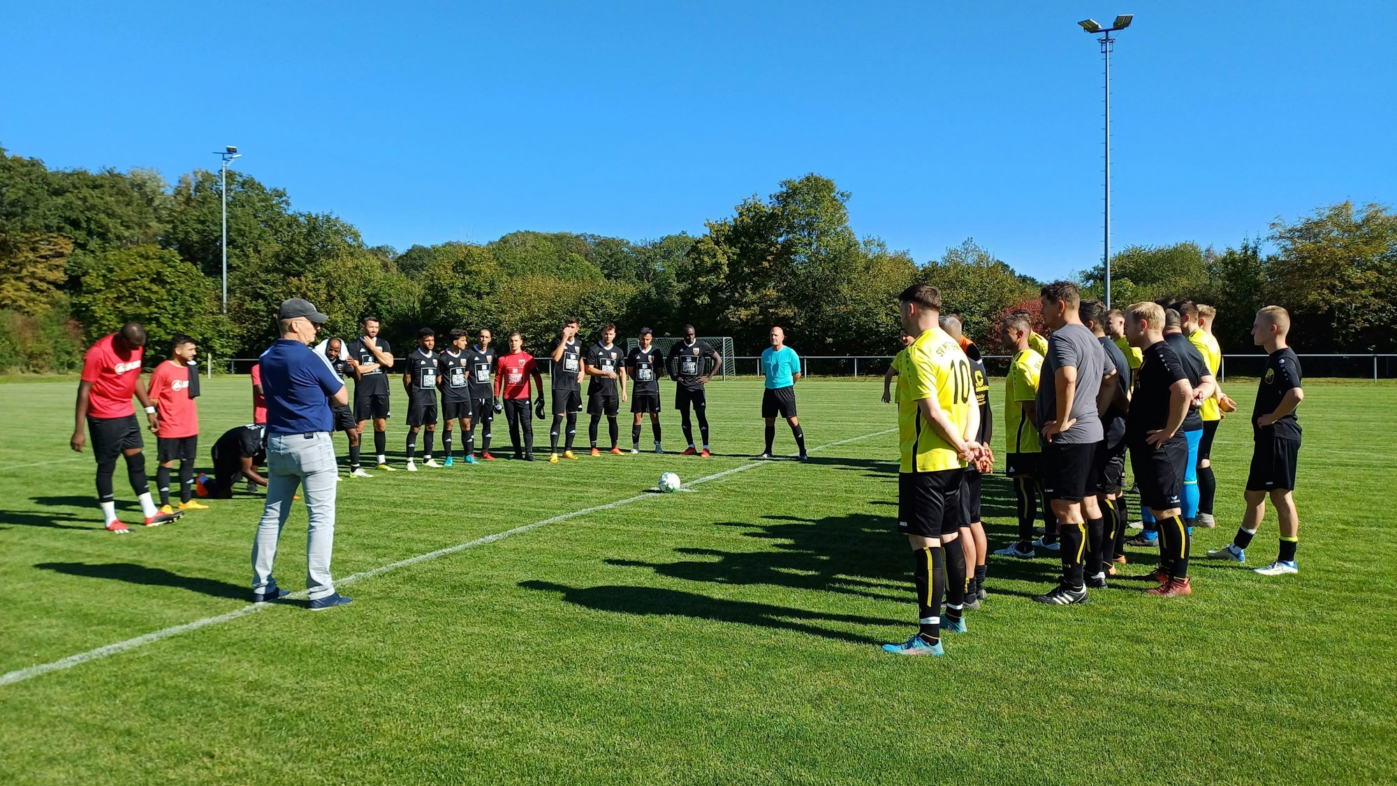 Ein Mann hält eine Ansprache, zwei Fußballteams stehen im Halbkreis um ihn herum und hören zu.