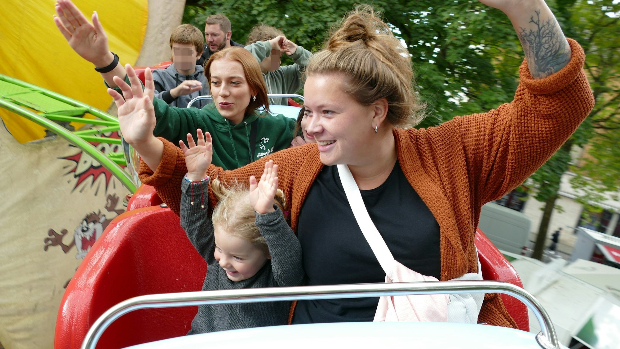 Ein kleines blondes Mädchen und seine Mutter sitzen in einem Achterbahnwagen und reißen die Arme hoch.