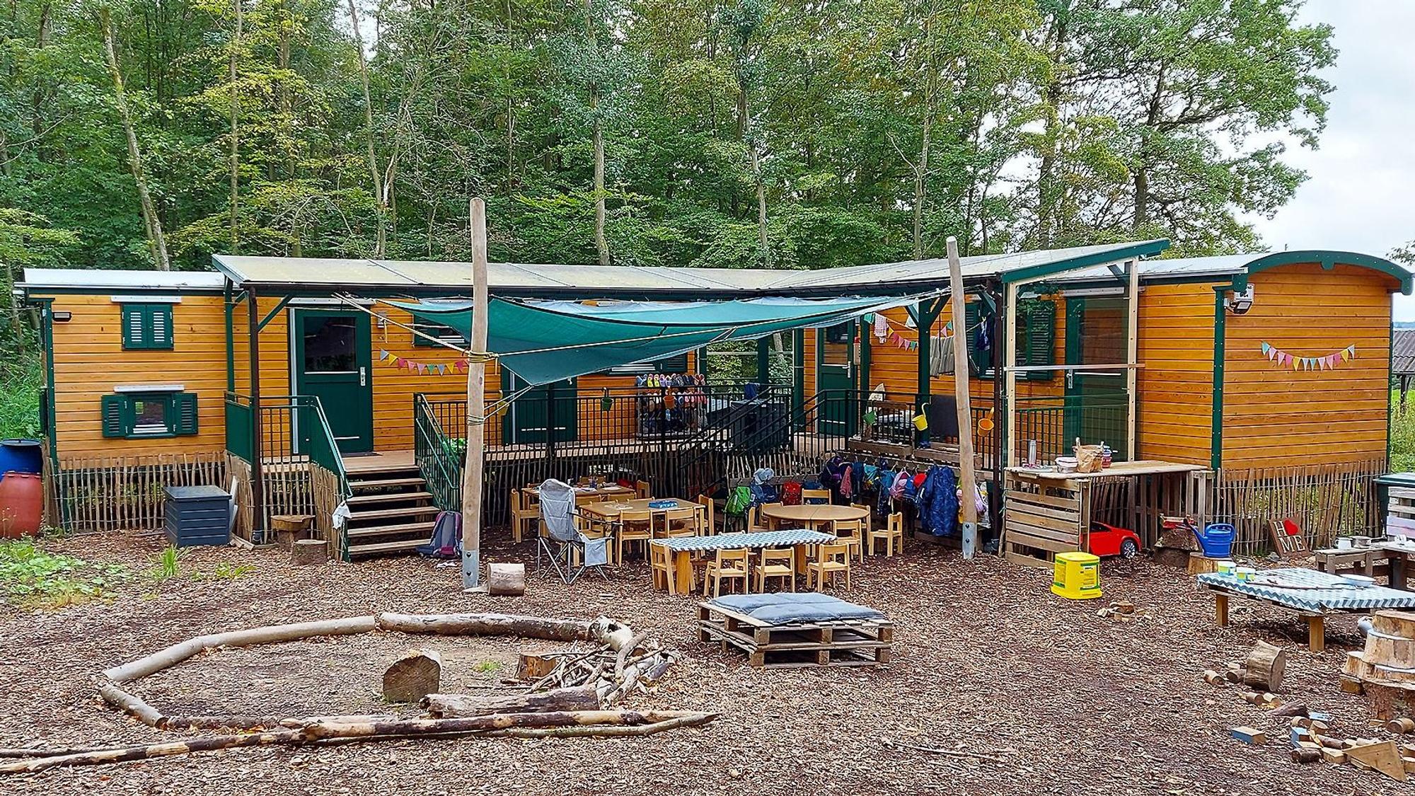 In Bauwagen aus Holz ist der Waldkindergarten Mülheim eingerichtet. Im Vordergrund sind Tische, Stühle und Spielsachen zu sehen, im Hintergrund Bäume.