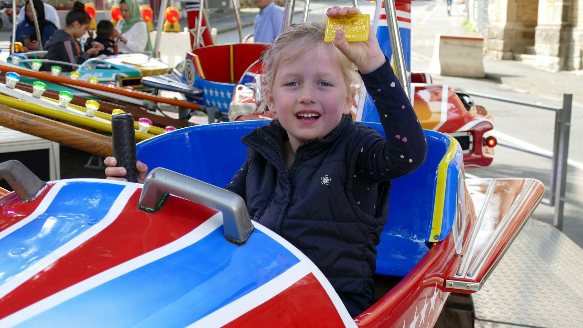 Ein Mädchen sitzt in einem rot-weißen Flugzeug in einem Kinderkarussell und hält einen Fahrchip in die Höhe.