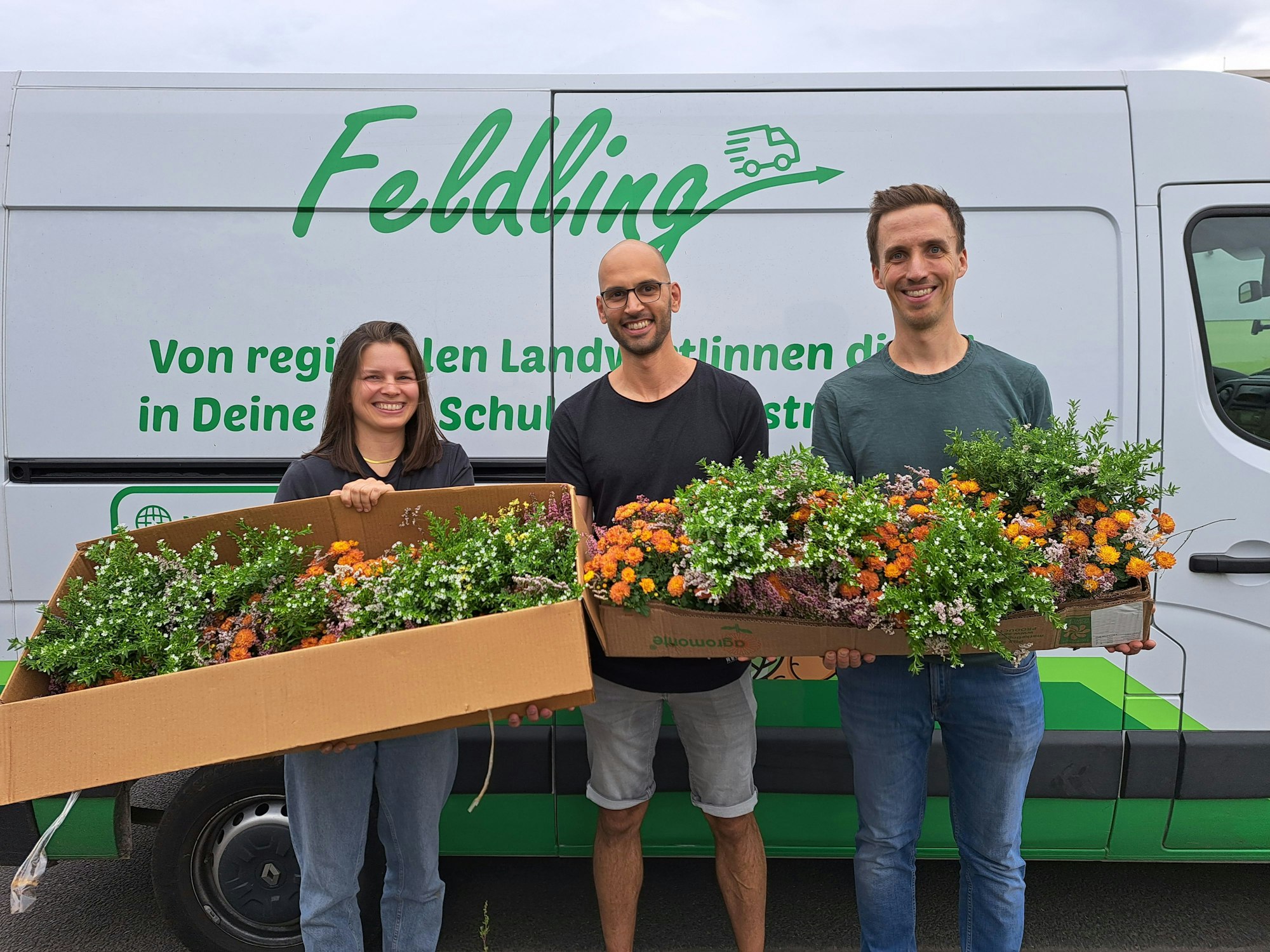 Eine Frau und zwei Männer stehen vor einem Transporter. Sie halten Kartons mit Blumen in den Händen.