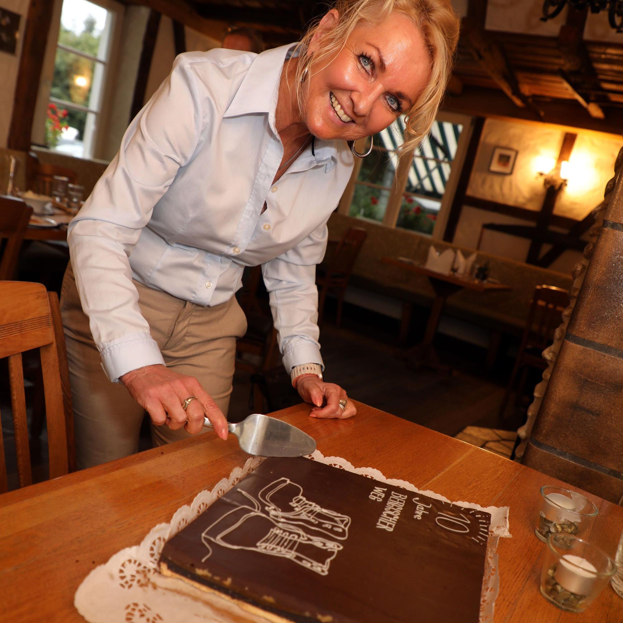 Gabi Wilhelm schneidet mit einer Kuchenschaufel einen Geburtstagstore an auf der „10 Jahre Bergischer Weg“ steht.