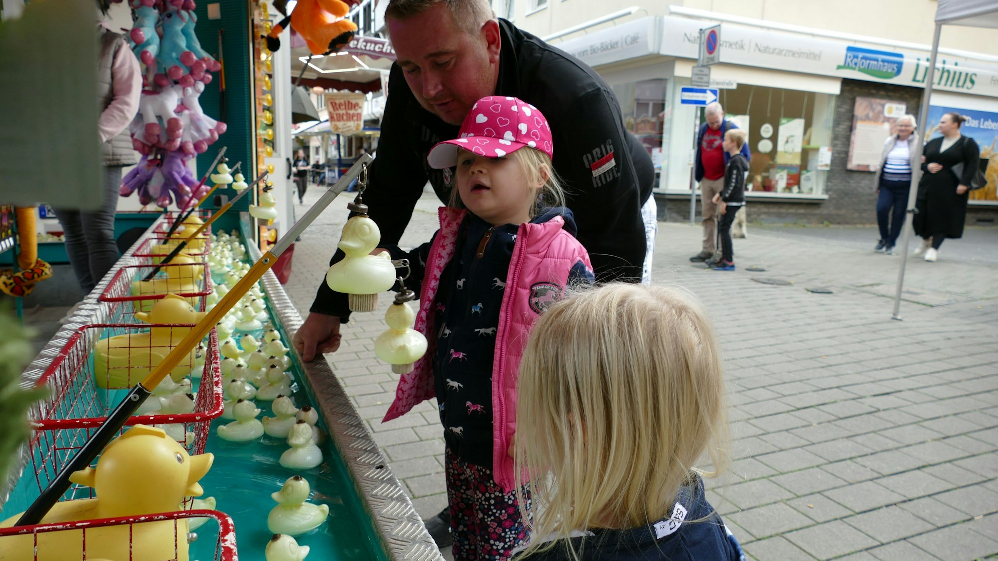 Ein Mann steht mit zwei kleinen blonden Mädchen vor einer Kirmesbude, in der Gummienten in einem Wasserbassin schwimmen.