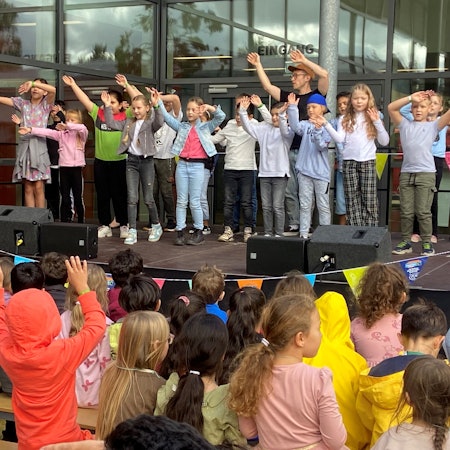 Kinder stehen mit erhobenen Armen auf einer Bühne und singen.