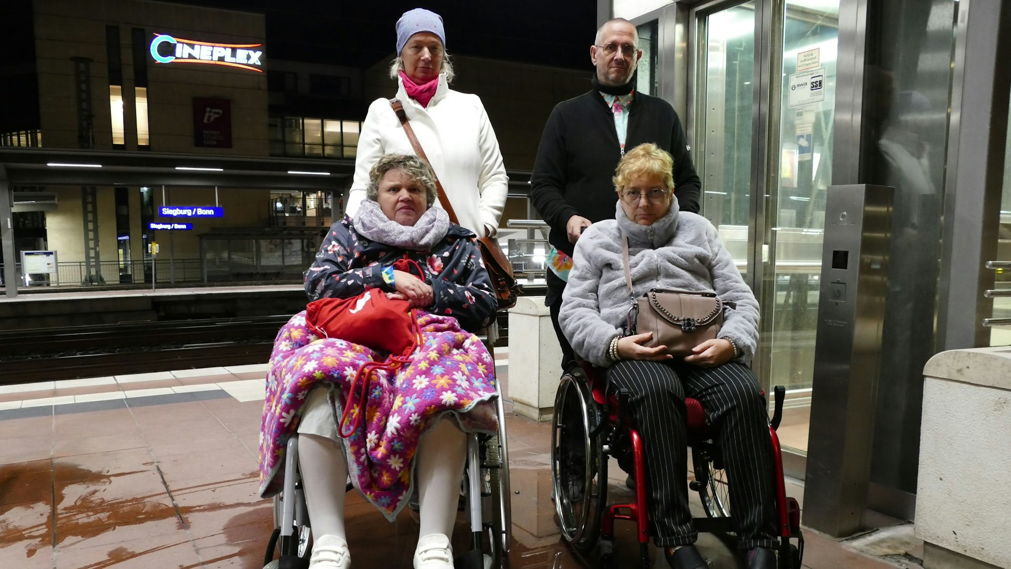 Zwei Frauen im Rollstuhl auf einem Bahnsteig, dahinter stehen eine Frau und ein Mann.
