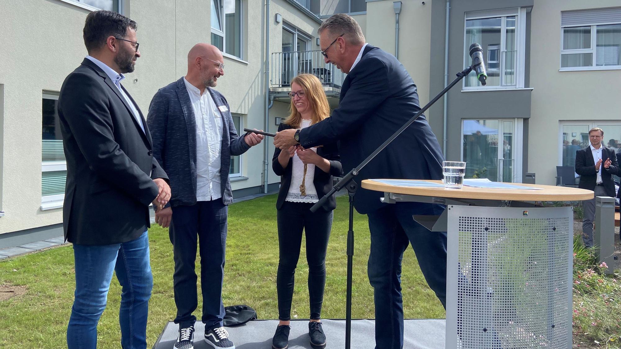 Drei Männer und eine Frau stehen vor einem Neubau neben einem Rednerpult. Einer der Männer übergibt einem anderen einen Schlüssel.