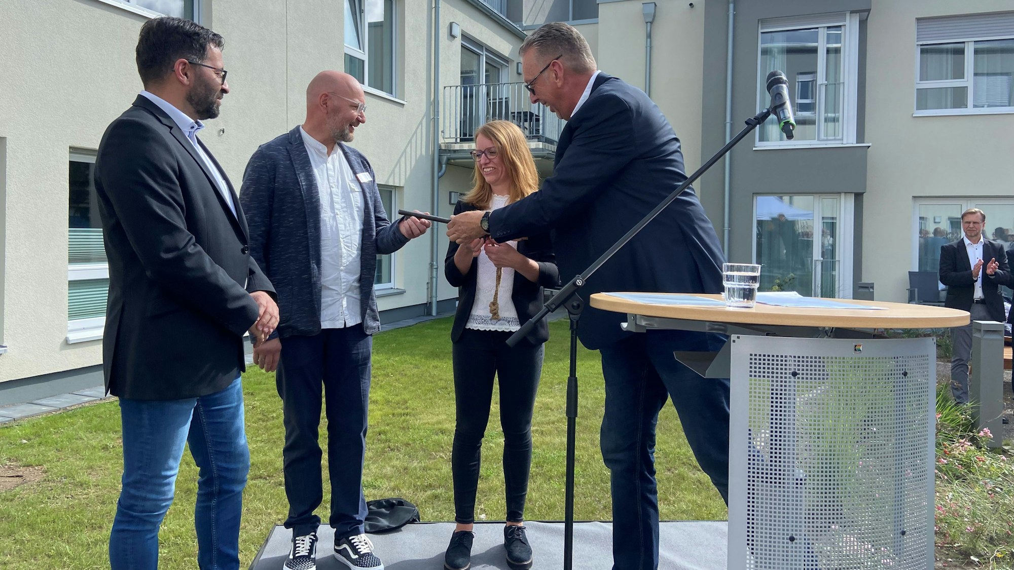 Drei Männer und eine Frau stehen vor einem Neubau neben einem Rednerpult. Einer der Männer übergibt einem anderen einen Schlüssel.
