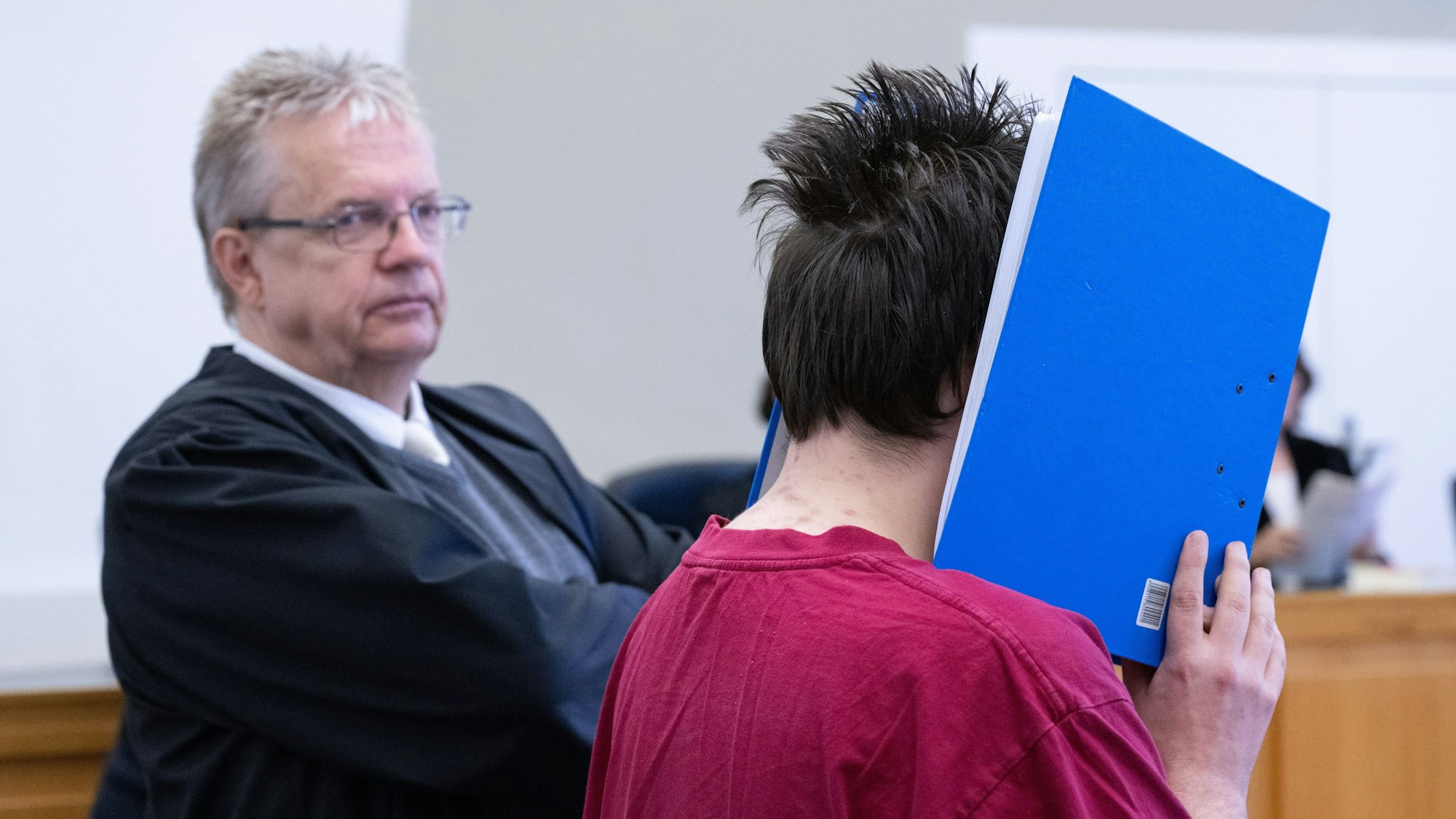 Der Angeklagte wird in den Verhandlungssaal geführt und verbirgt sein Gesicht hinter einem Aktenordner. Daneben steht sein Anwalt Henner Maaß.