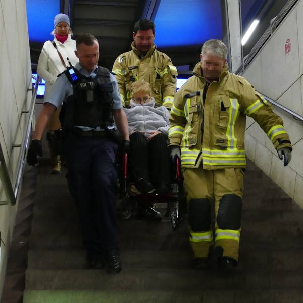 Zwei Feuerwehrmänner in gelben Overalls und ein Bundespolizist in blau tragen eine Frau im Rollstuhl eine Treppe heyrunter.