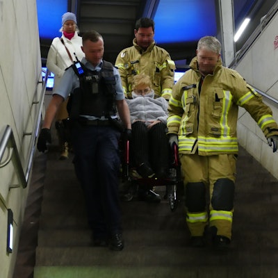 Zwei Feuerwehrmänner in gelben Overalls und ein Bundespolizist in blau tragen eine Frau im Rollstuhl eine Treppe heyrunter.