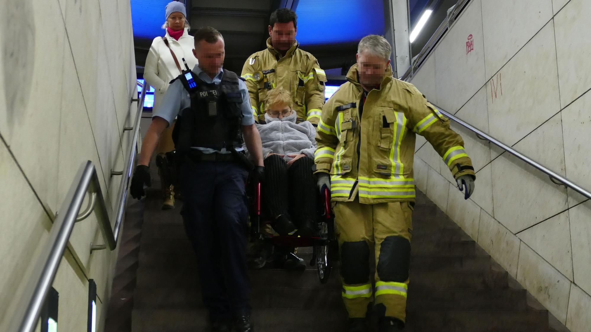 Zwei Feuerwehrmänner in gelben Overalls und ein Bundespolizist in blau tragen eine Frau im Rollstuhl eine Treppe heyrunter.