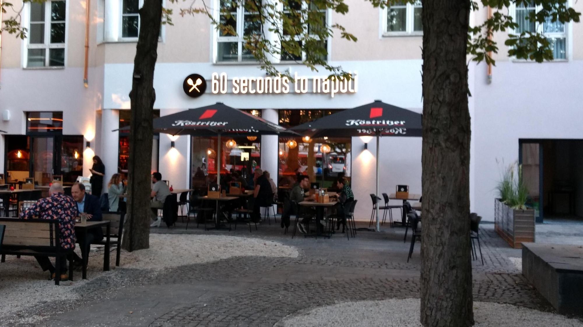 Vor einem Gebäude stehen Tische, Stühle und Sonnenschirme. An der Fasse steht eine Aufschrift mit „60 seconds to napoli“.