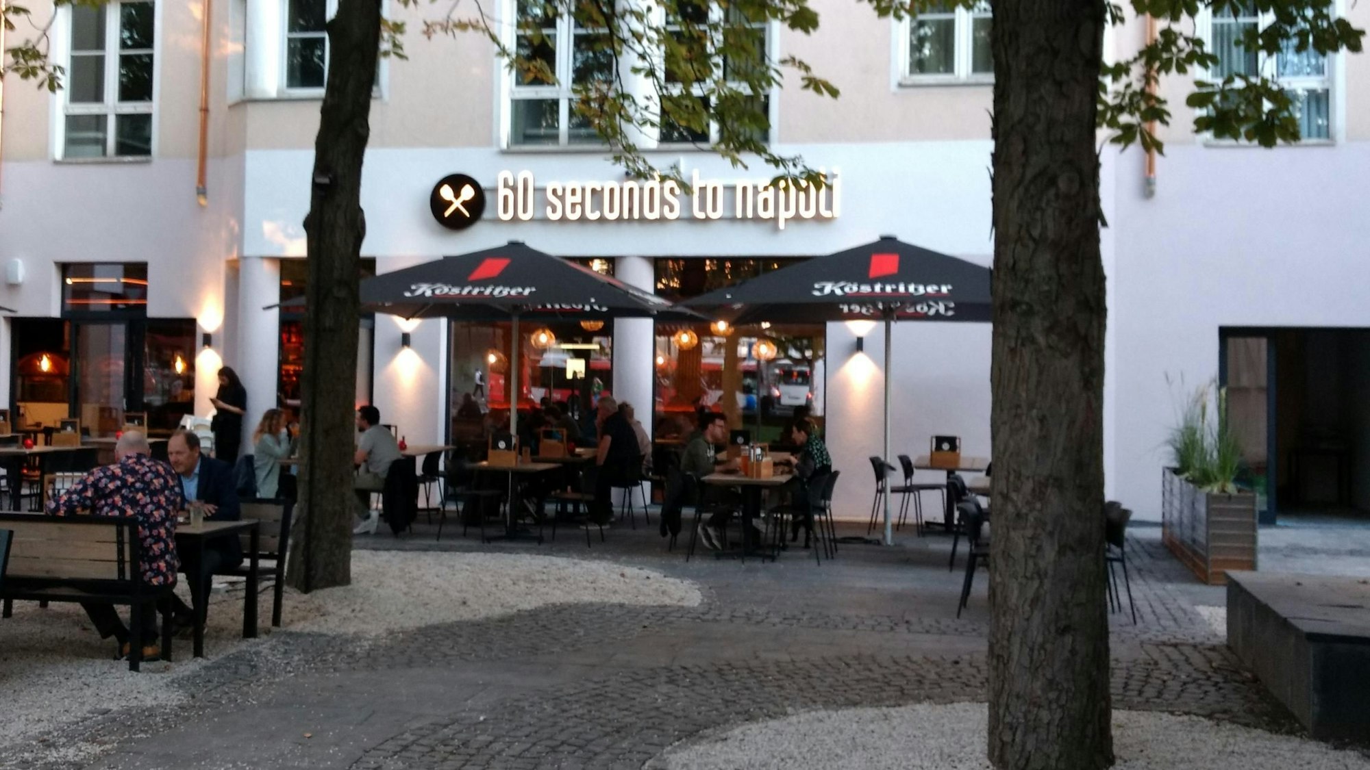 Vor einem Gebäude stehen Tische, Stühle und Sonnenschirme. An der Fasse steht eine Aufschrift mit „60 seconds to napoli“.