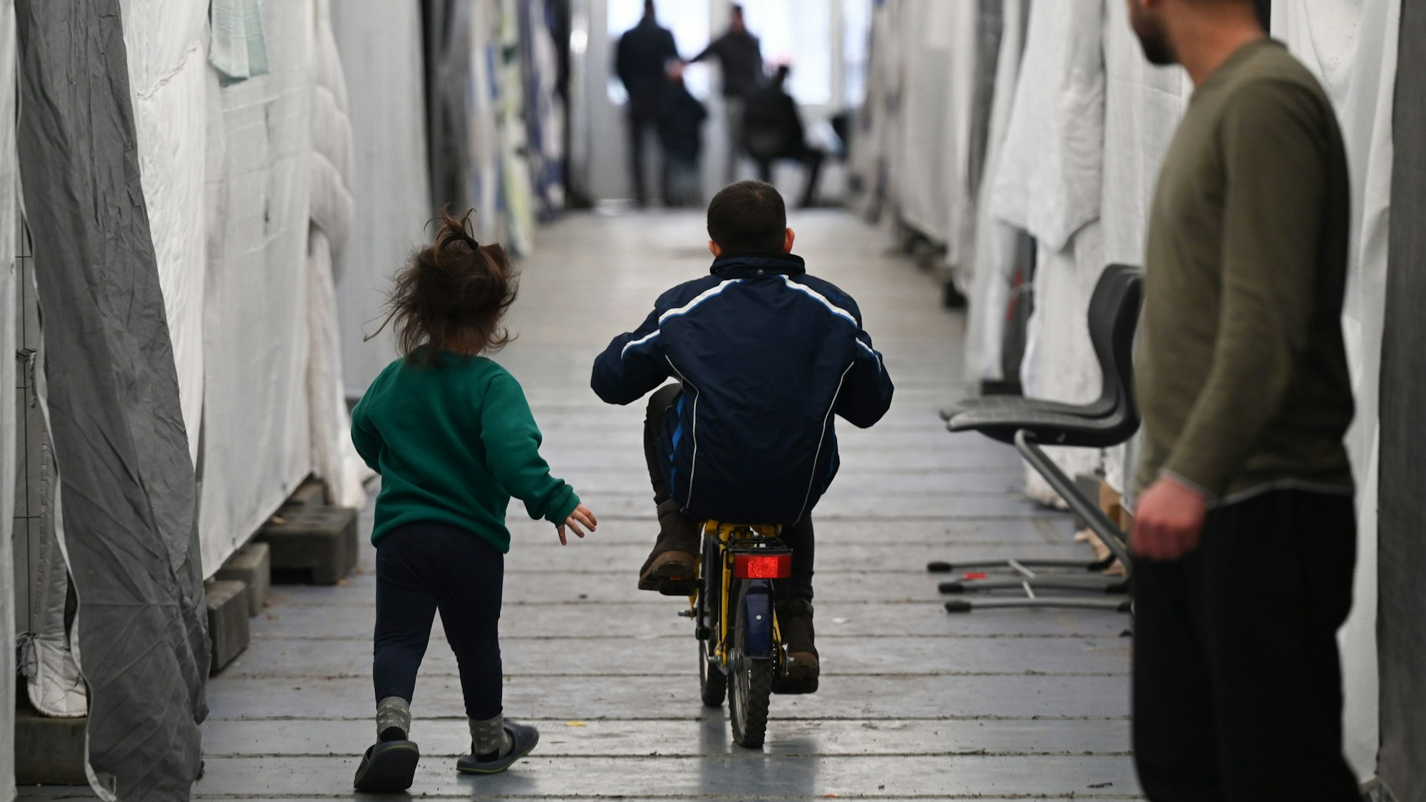 Flüchtlinge in Zelten, Wohncontainern und Turnhallen gehören auch in NRW längst wieder zur Realität. Ein kleiner Junge fährt auf einem Fahrrad zwischen Planen hindurch, ein kleines Mädchen eilt hinter ihm her.