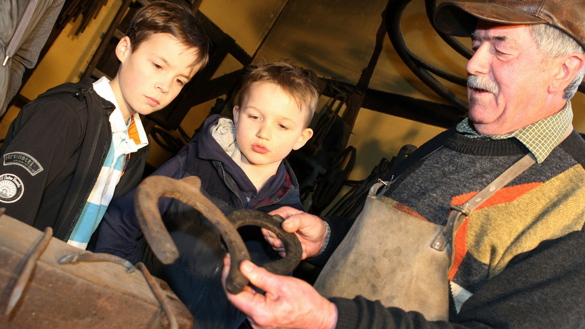 Ein Schmied zeigt zwei Kindern Hufeisen in der Schmiede.
