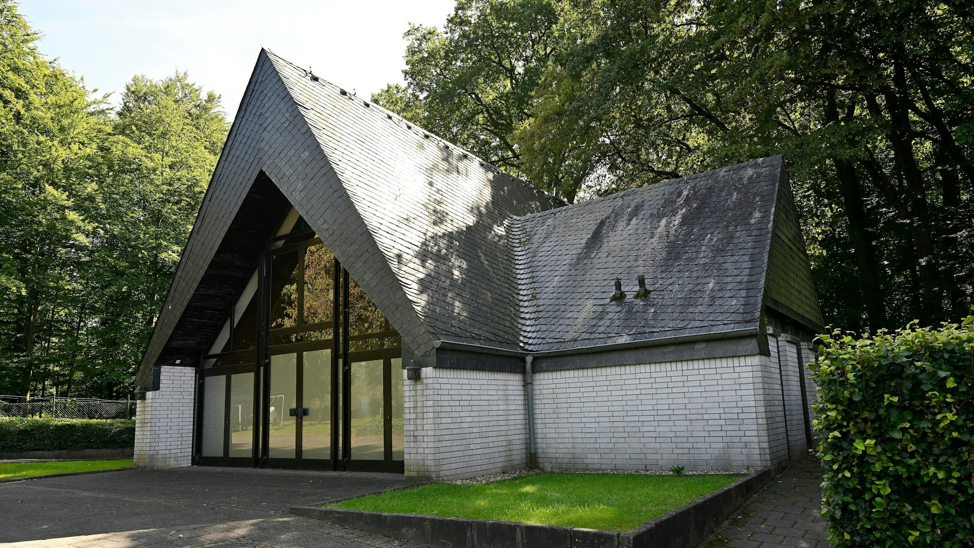 Eine Trauerhalle ist von außen zu sehen: ein steil sich zuspitzendes schwarzes Dach auf weißen Grundmauern mit einem großen Glasportal. Im Hintergrund stehen Bäume.