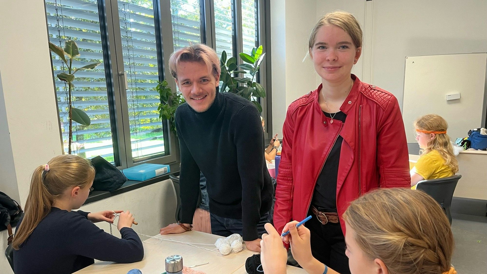 In einem Klassenraum stehen zwei Freiwillige an einem Tisch mit Schülerinnen.