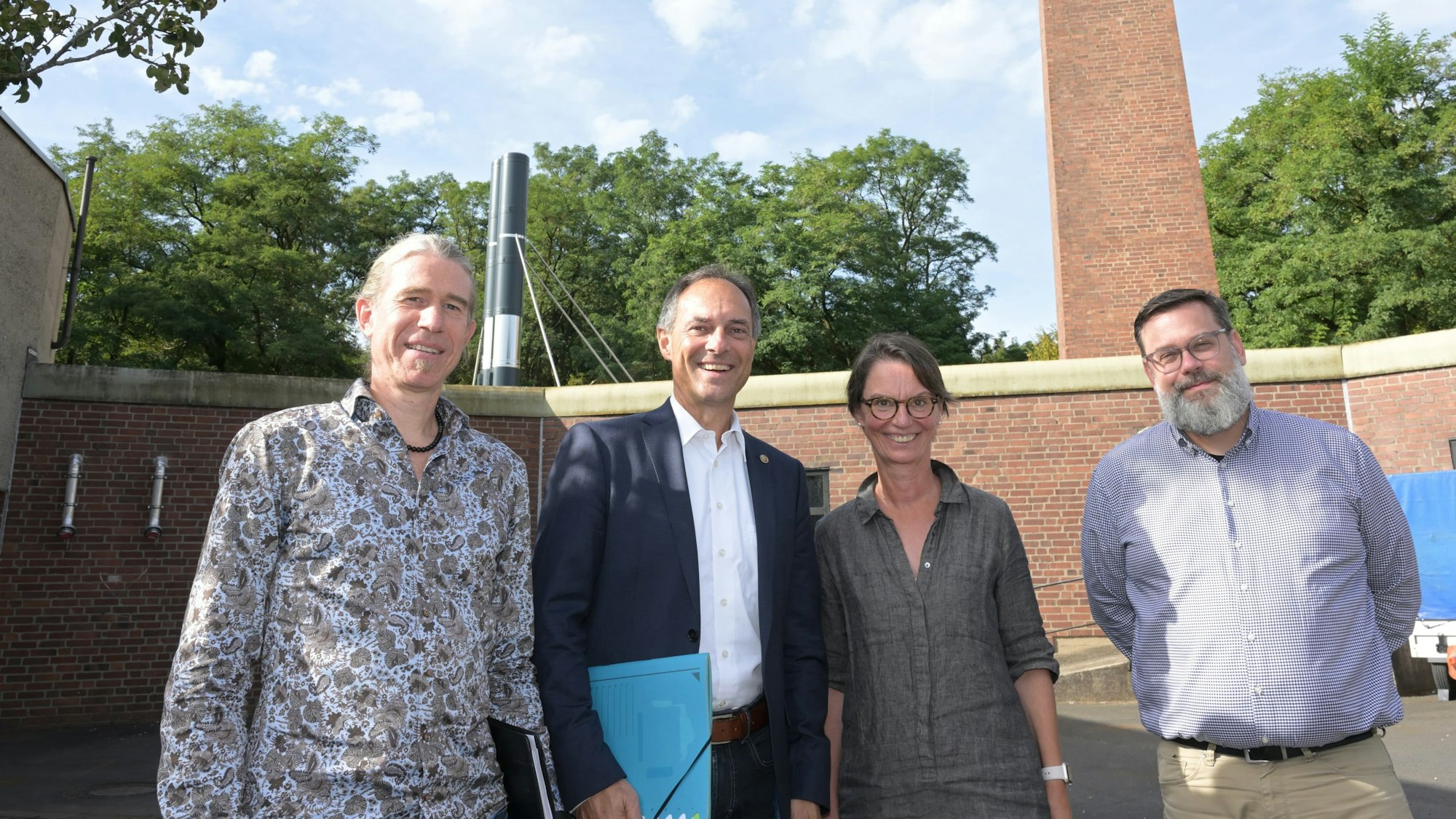 Marco Preußer, Manfred Habrunner, Jutta Menne, Dominik Haupt stehen nebeneinander vor der Heizungszentrale, einem Backsteinbau mit einem hohen Kamin.