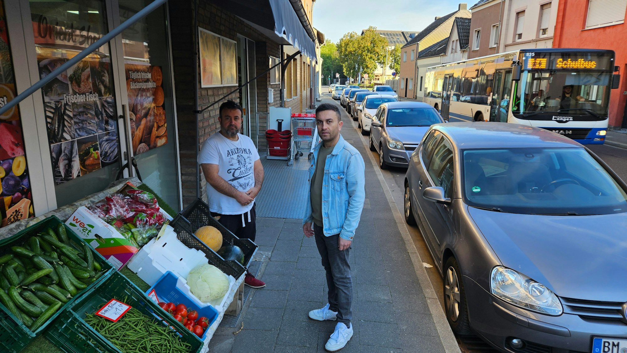 Zwei Männer stehen vor einem Mini-Supermarkt mit Gemüseauslage an einer verkehrsreichen Straße.