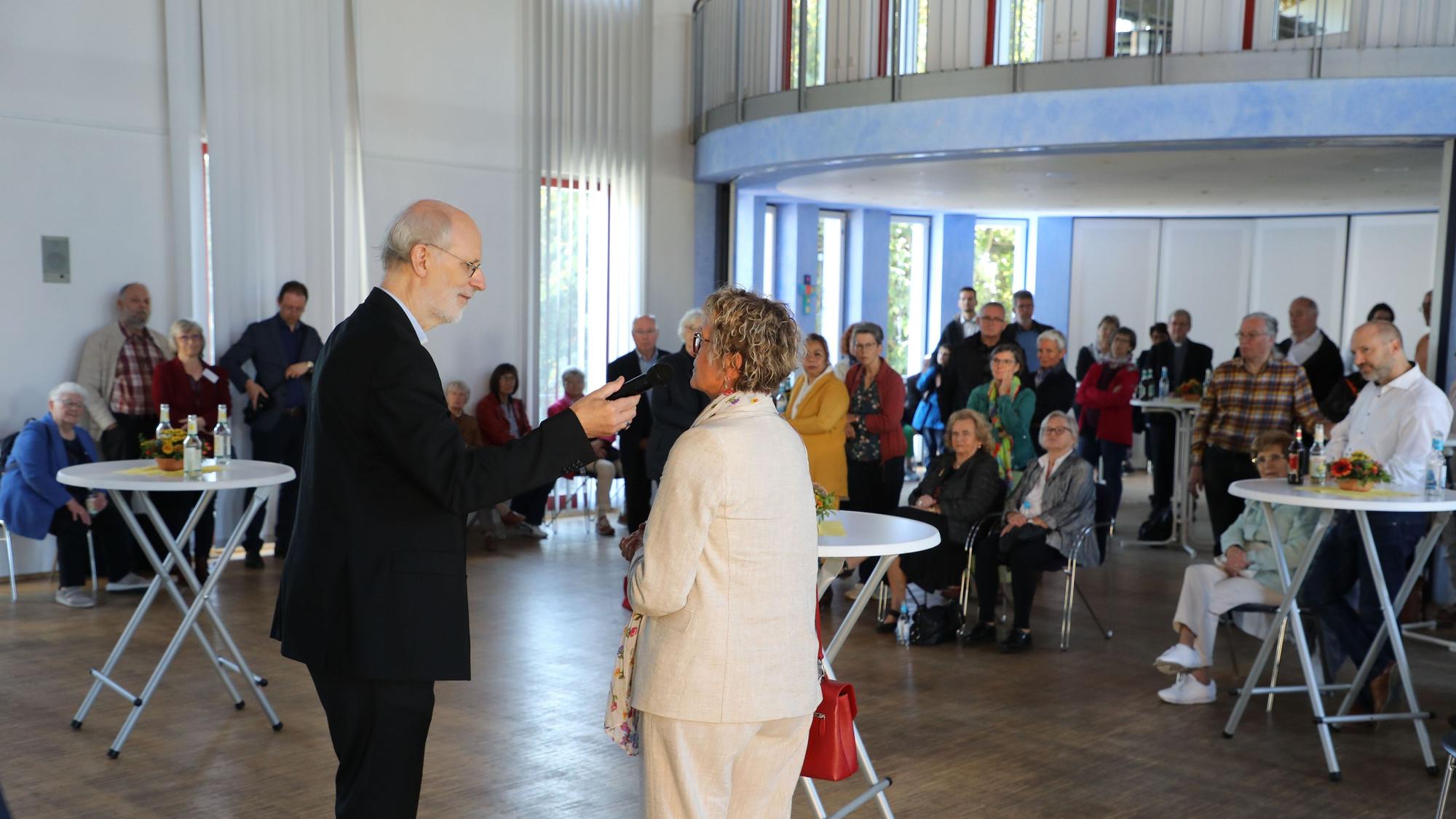 Zwei Menschen mit Mikrofon stehen vor einer Versammlung.
