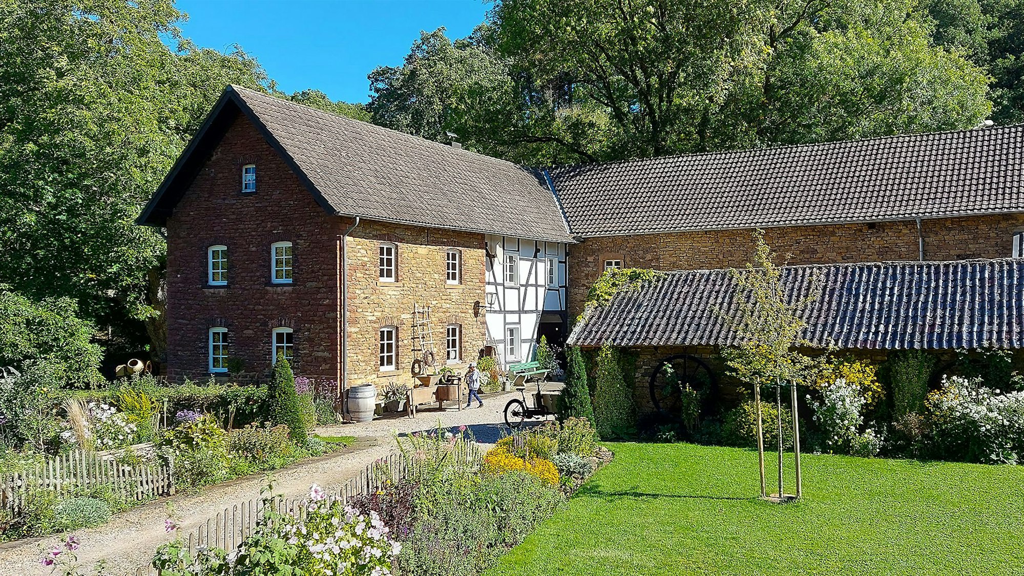 Die Mühle Falkenstein in Eiserfey, umgeben von einem Staudengarten, Wald und einer Wiese.