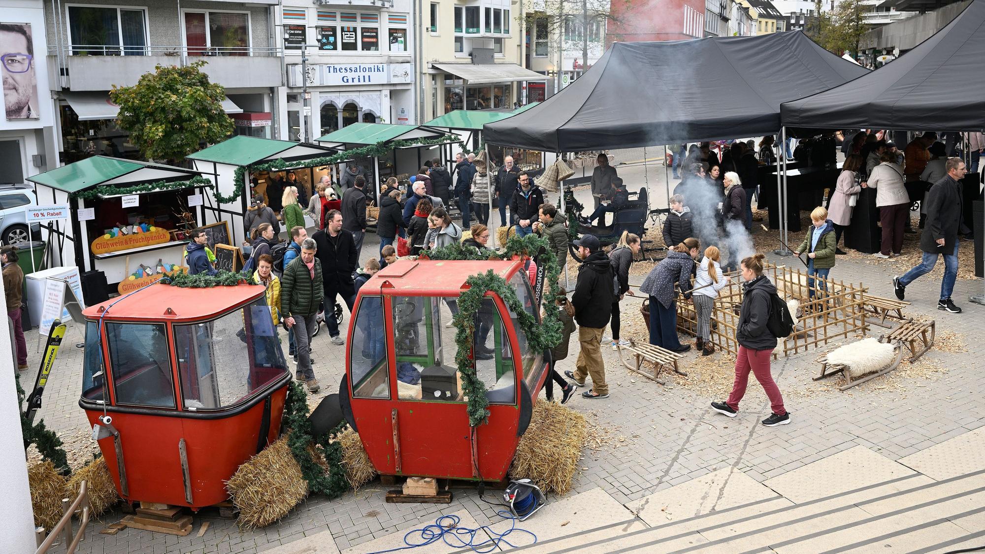 Auf einem Platz tummeln sich Menschen und Pavillon-Zelten und an Marktständen. Um eine Feuerstelle sind Sitzgelegenheiten aufgestellt, darunter auch alte Seilbahnwagen.
