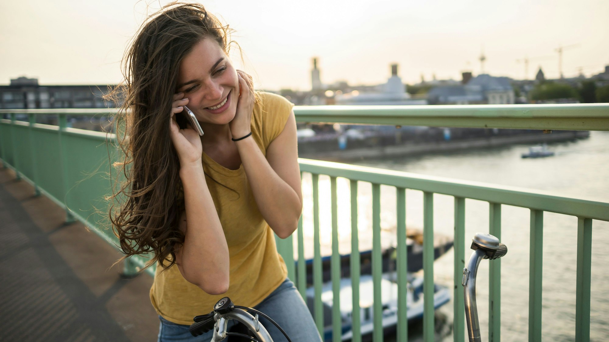 Eine Frau steht auf einer Brücke in Köln auf ihrem stehenden Fahrrad und telefoniert