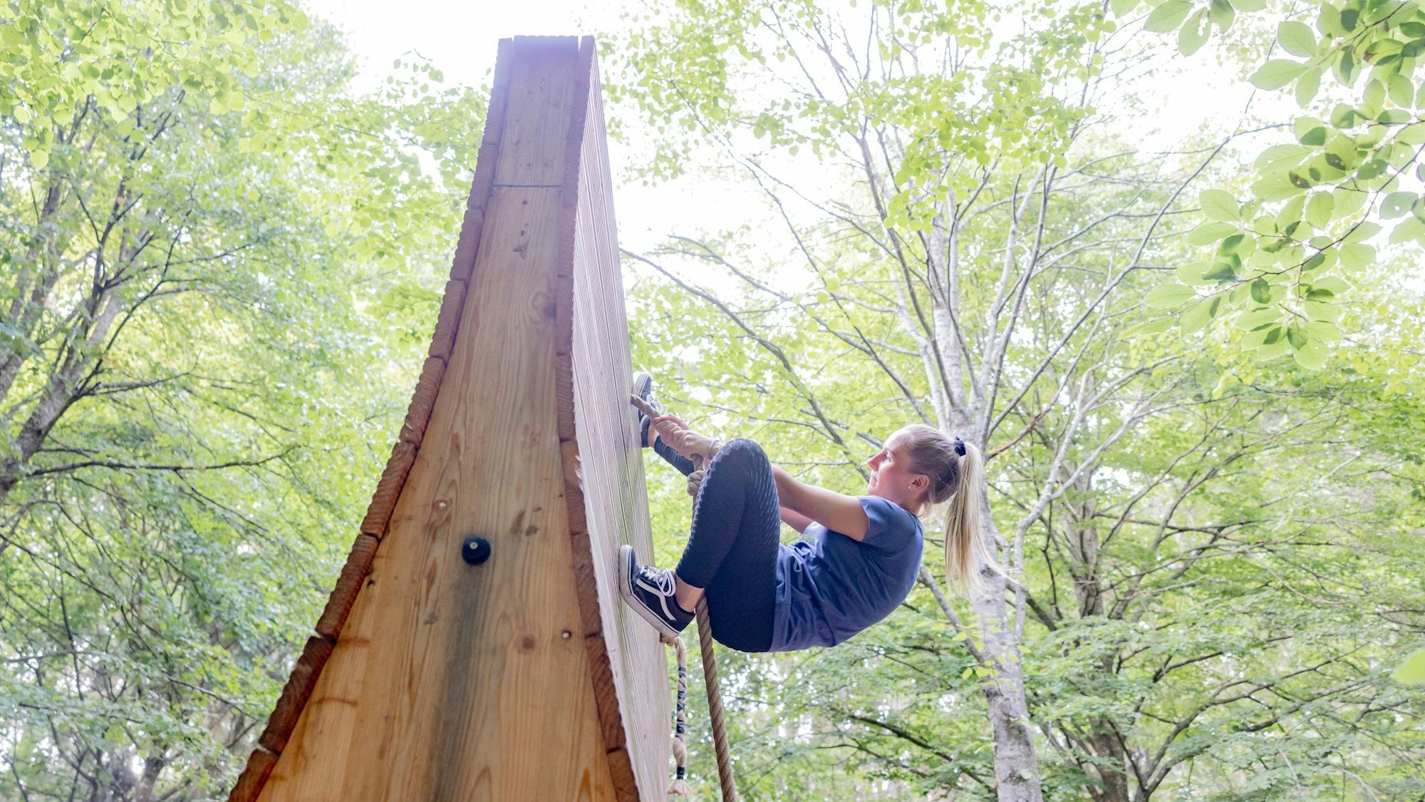eine Frau klettert in einem Wald eine Wand aus Holz an einem Seil hoch