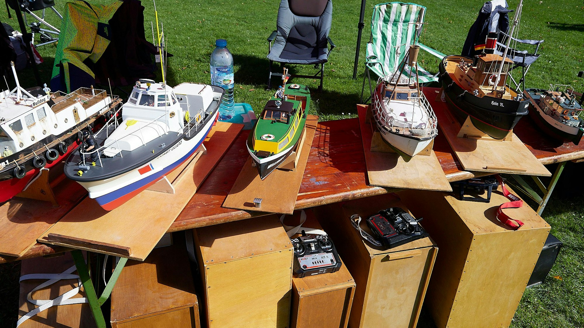 Auf einem Tisch stehen zahlreiche Modellboote. Die Fernsteuerungen dafür sind auf Kisten darunter abgelegt.