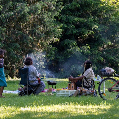 Zwei Menschen sitzen auf einer Wiese und grillen, eine Frau steht daneben.