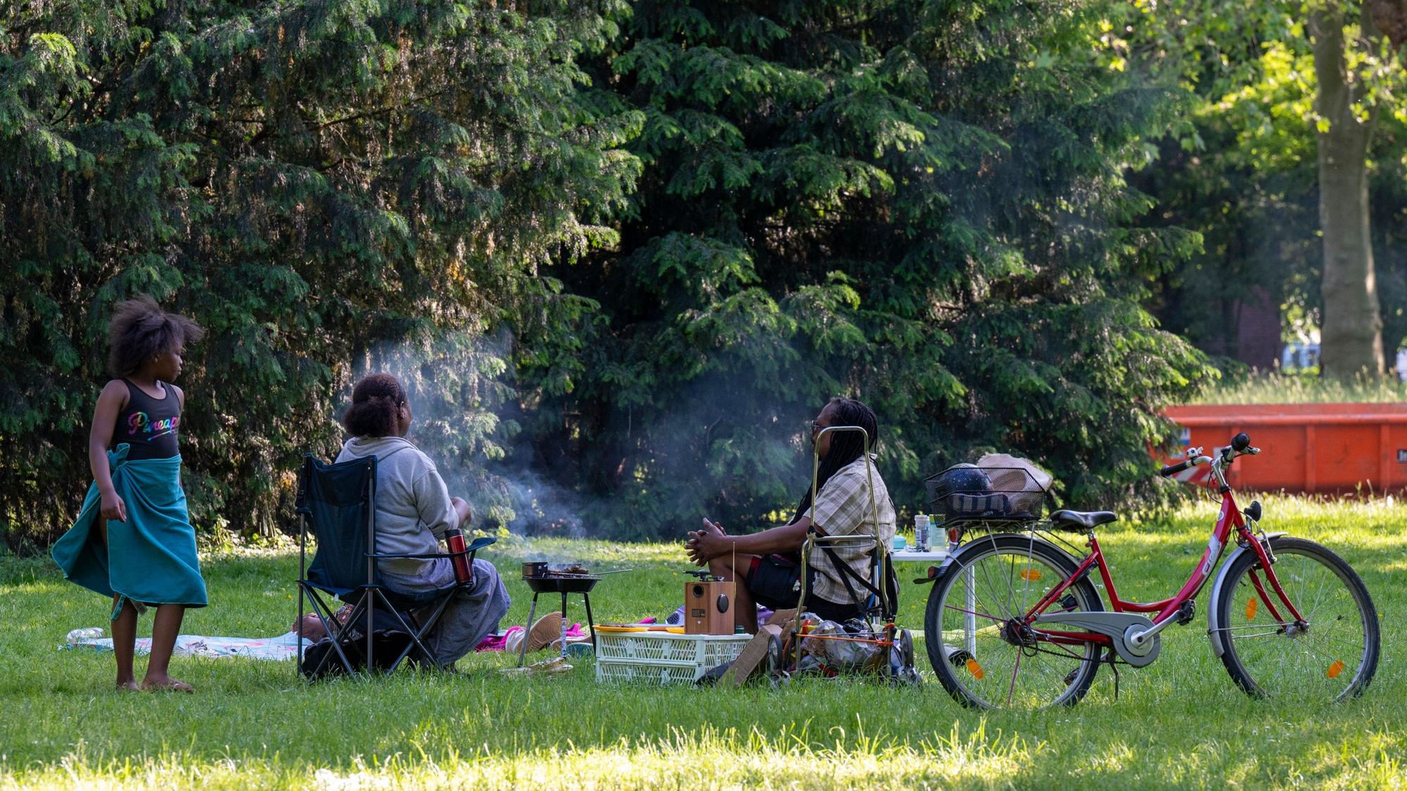 Zwei Menschen sitzen auf einer Wiese und grillen, eine Frau steht daneben.