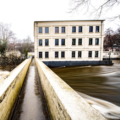 Das Foto zeigt die Fußgängerbrücke über die Wupper mit dem Alten Turbinenhaus und dem Wehr im Hintergrund.