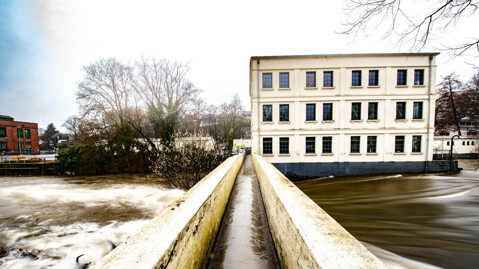 Das Foto zeigt die Fußgängerbrücke über die Wupper mit dem Alten Turbinenhaus und dem Wehr im Hintergrund.