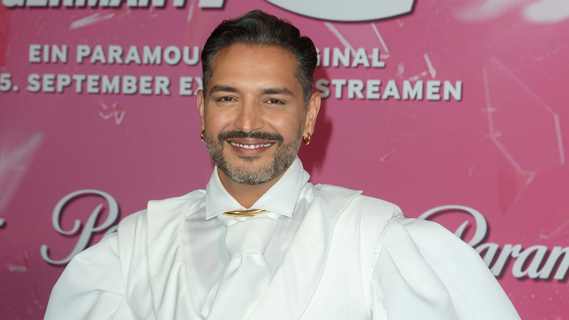 Gianni Jovanovic bei der Premiere der Show „Drag Race Germany“, die beim Streamingdienst Paramount+ zu sehen ist. Er trägt ein weißes Hemd, eine weiße Krawatte und lächelt in die Kamera.