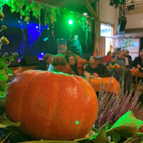 Im Vordergrund liegt ein Kürbis auf einem Bett aus bunten Pflanzen. Im Hintergrund sitzen Gruppen von Menschen an Tischen in einem Saal und essen und trinken.