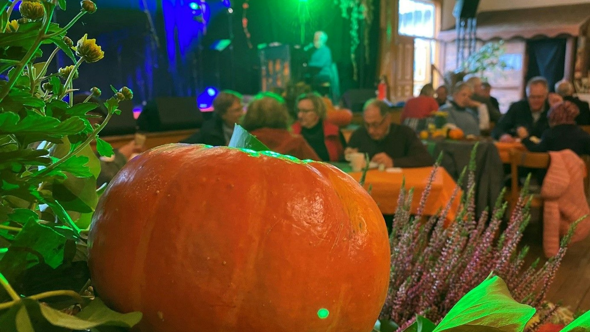 Im Vordergrund liegt ein Kürbis auf einem Bett aus bunten Pflanzen. Im Hintergrund sitzen Gruppen von Menschen an Tischen in einem Saal und essen und trinken.