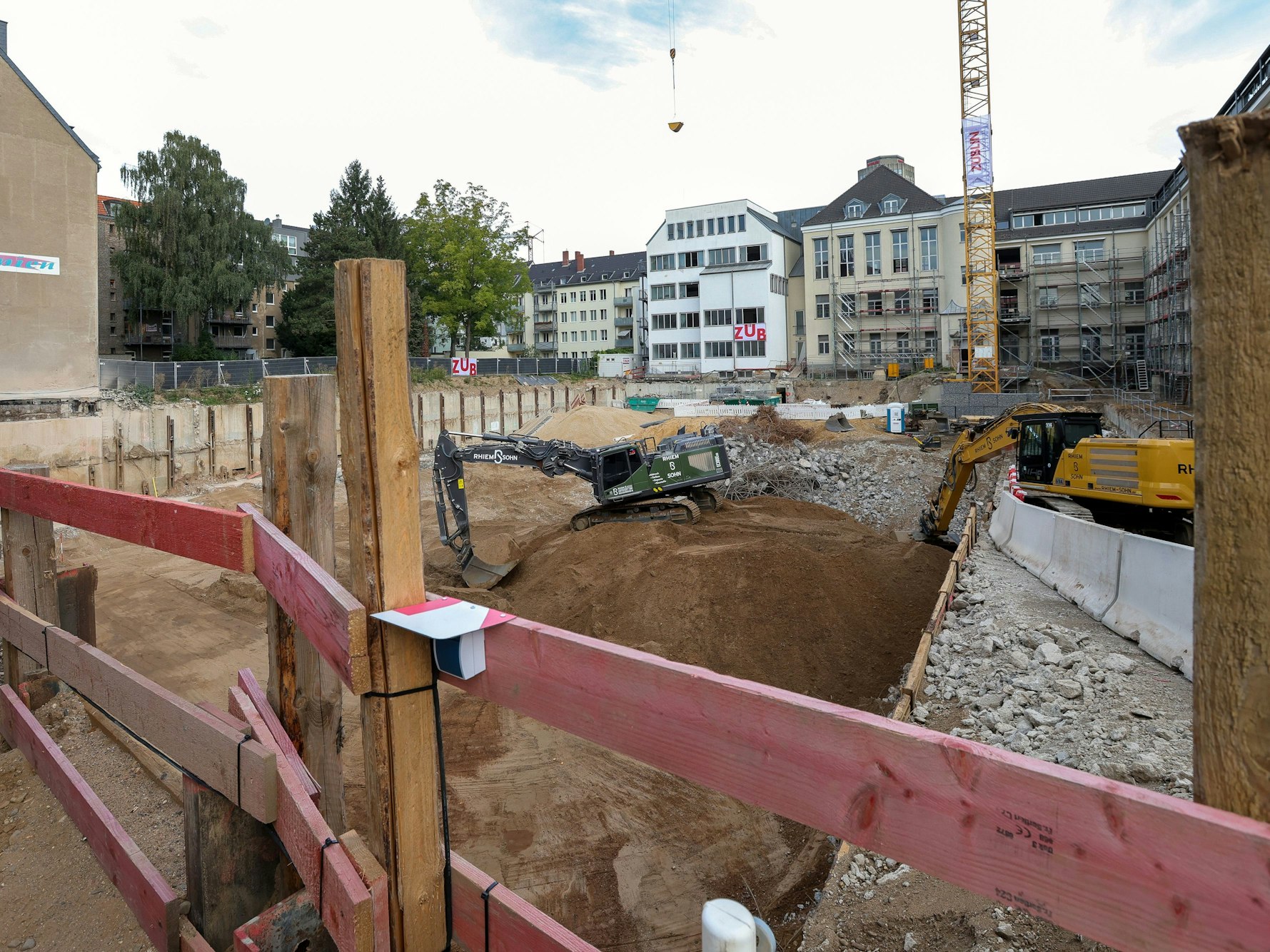 Ein Zaun aus Holzbrettern, dahinter eine große Baulücke mit Baumaschinen