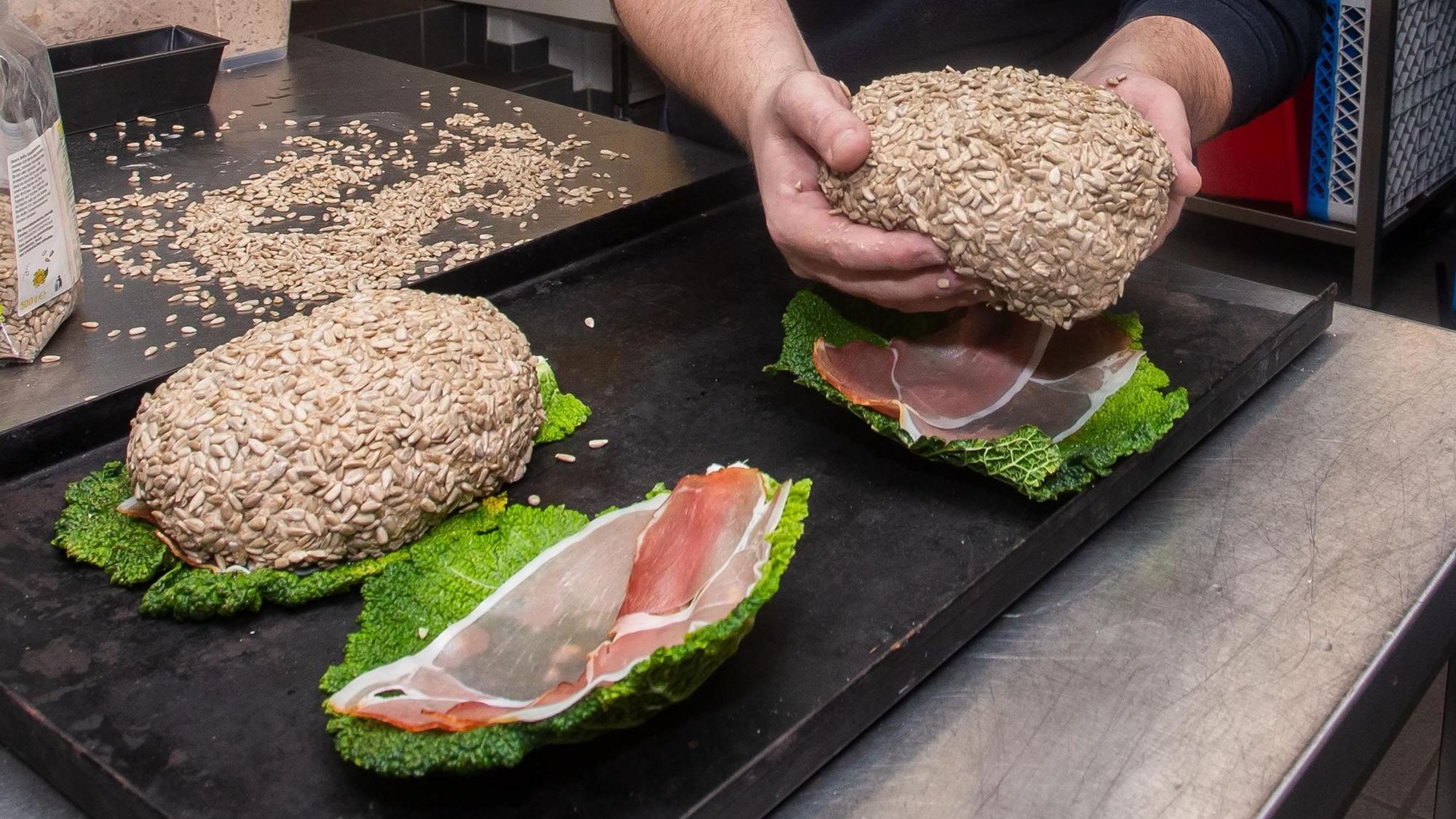 Ein Mann hält ein Brot in der Hand, das er auf ein Bett aus Salatblatt und rohem Schinken legt, bevor es in den Backofen kommt.