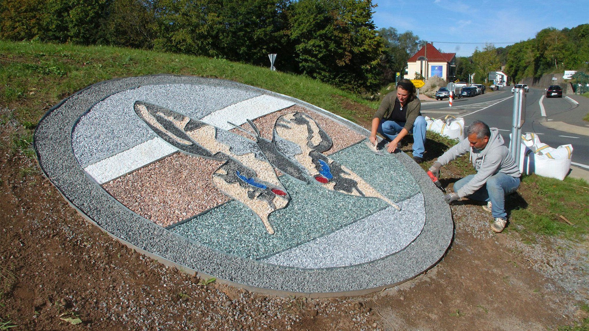 Auf der Insel des Kreisverkehrs in Ahrhütte legen ein Mann und eine Frau ein Schmetterlings-Bild aus farbigem Splitt.