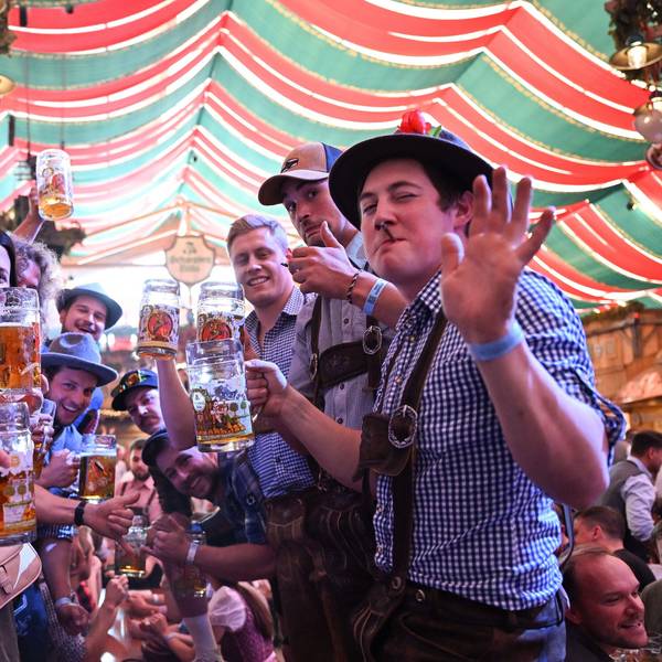 Junge Männer im blau-karierten Hemden und Lederhosen stehen in einer Art Spalier in einem Zelt und heben ihre Maßkrüge.