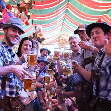 Junge Männer im blau-karierten Hemden und Lederhosen stehen in einer Art Spalier in einem Zelt und heben ihre Maßkrüge.