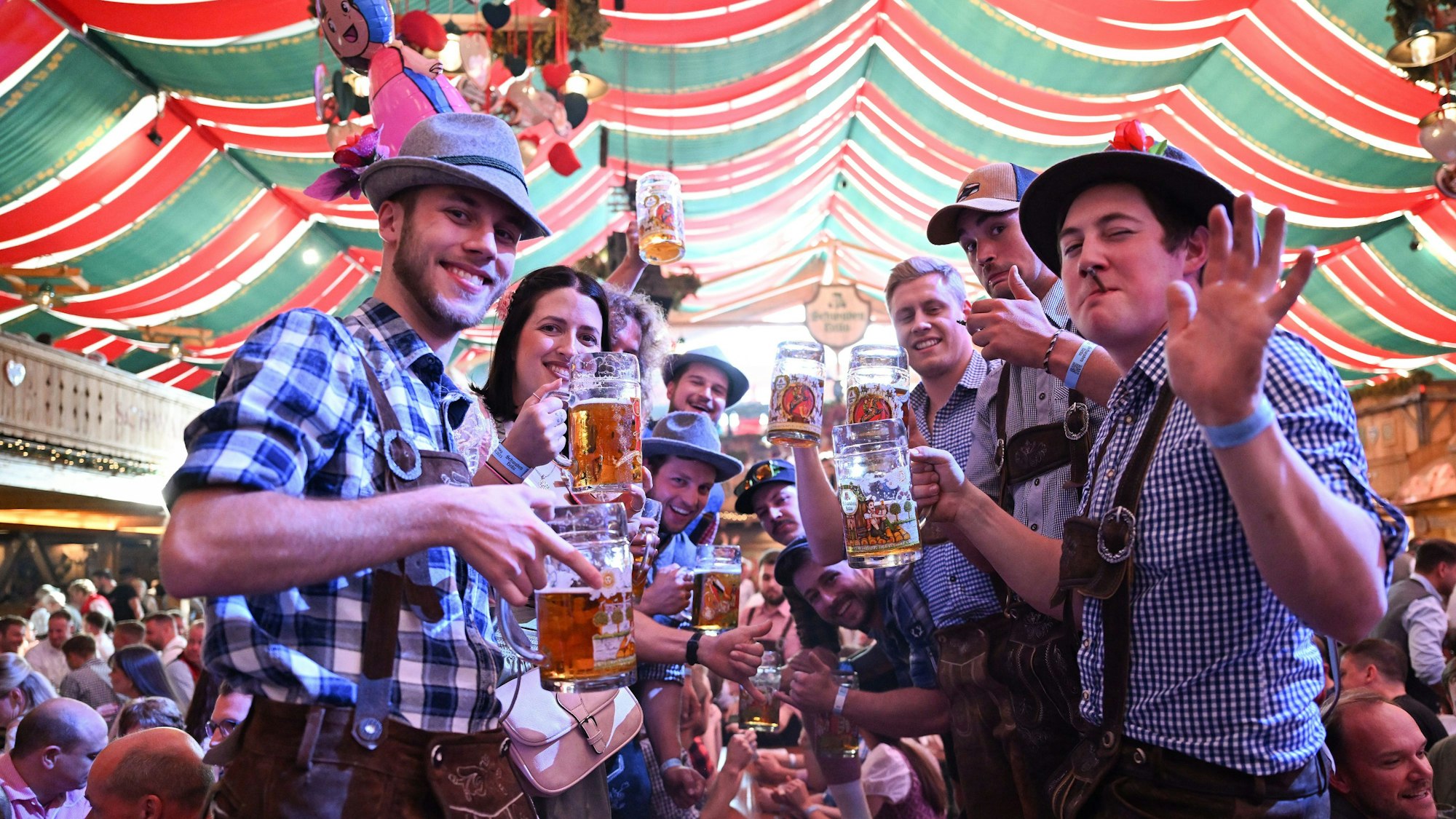 Junge Männer im blau-karierten Hemden und Lederhosen stehen in einer Art Spalier in einem Zelt und heben ihre Maßkrüge.