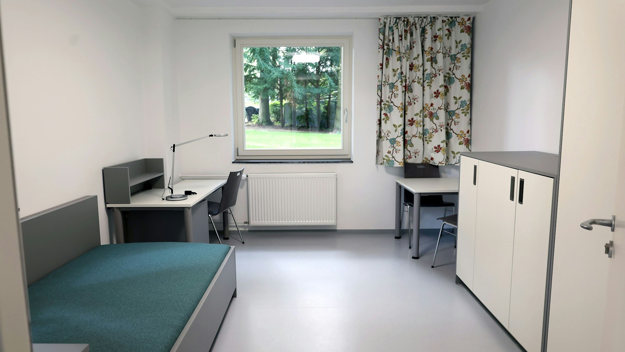 Schreibtisch, Kommode, kleiner Tisch, und Bett sowie ein Schrnak befinden sich in dem Zimmer.