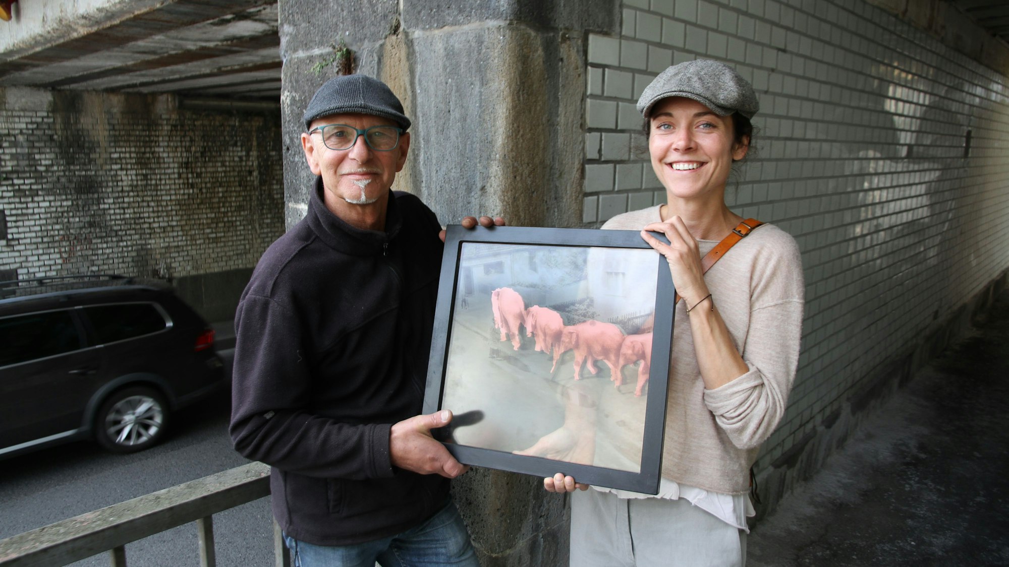 Ein Künstler und eine Künstlerin stehen mit einem Bild, das rosafarbene Elefanten zeigt, an einer Bahnunterführung an der Mahrstraße