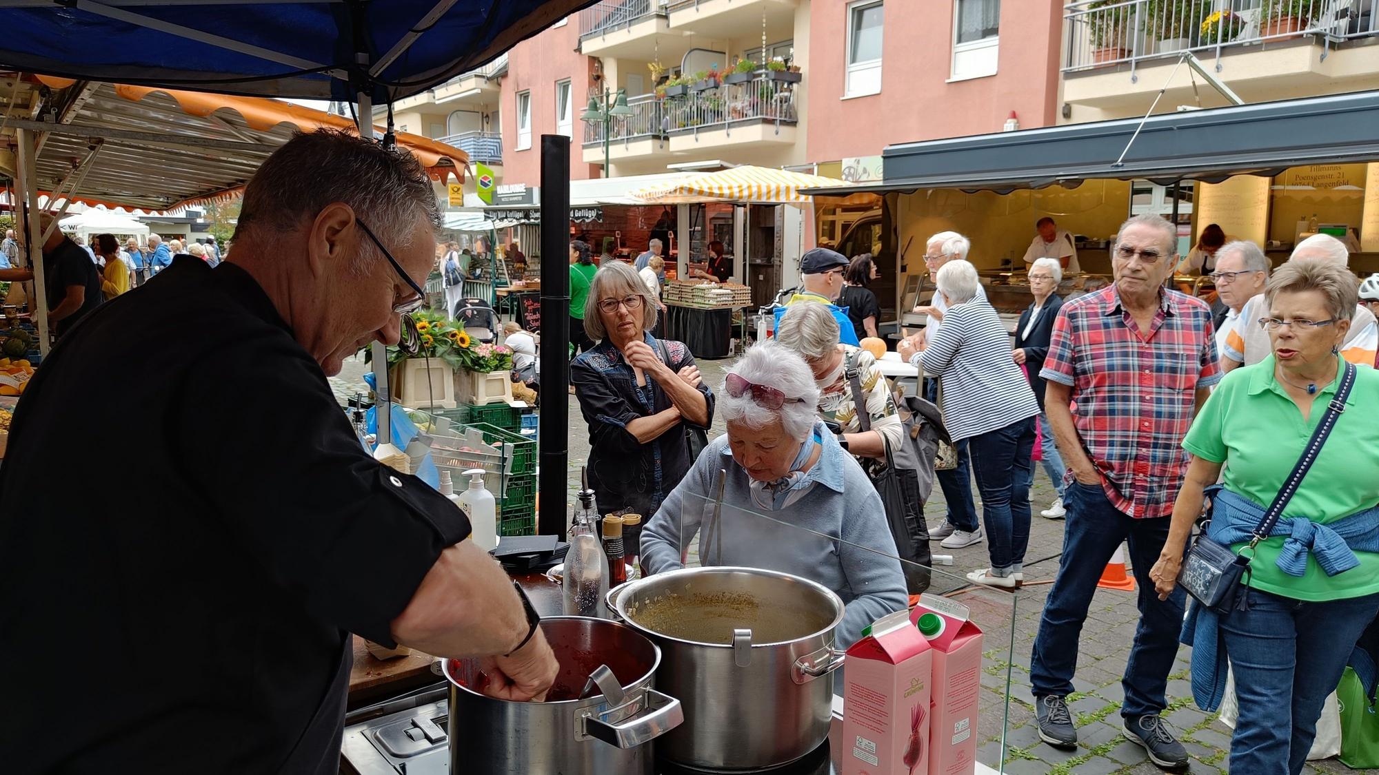 Ein Mann steht an einem Herd auf einem Marktplatz. Vor dem Herd Besucherinnen und Besucher.