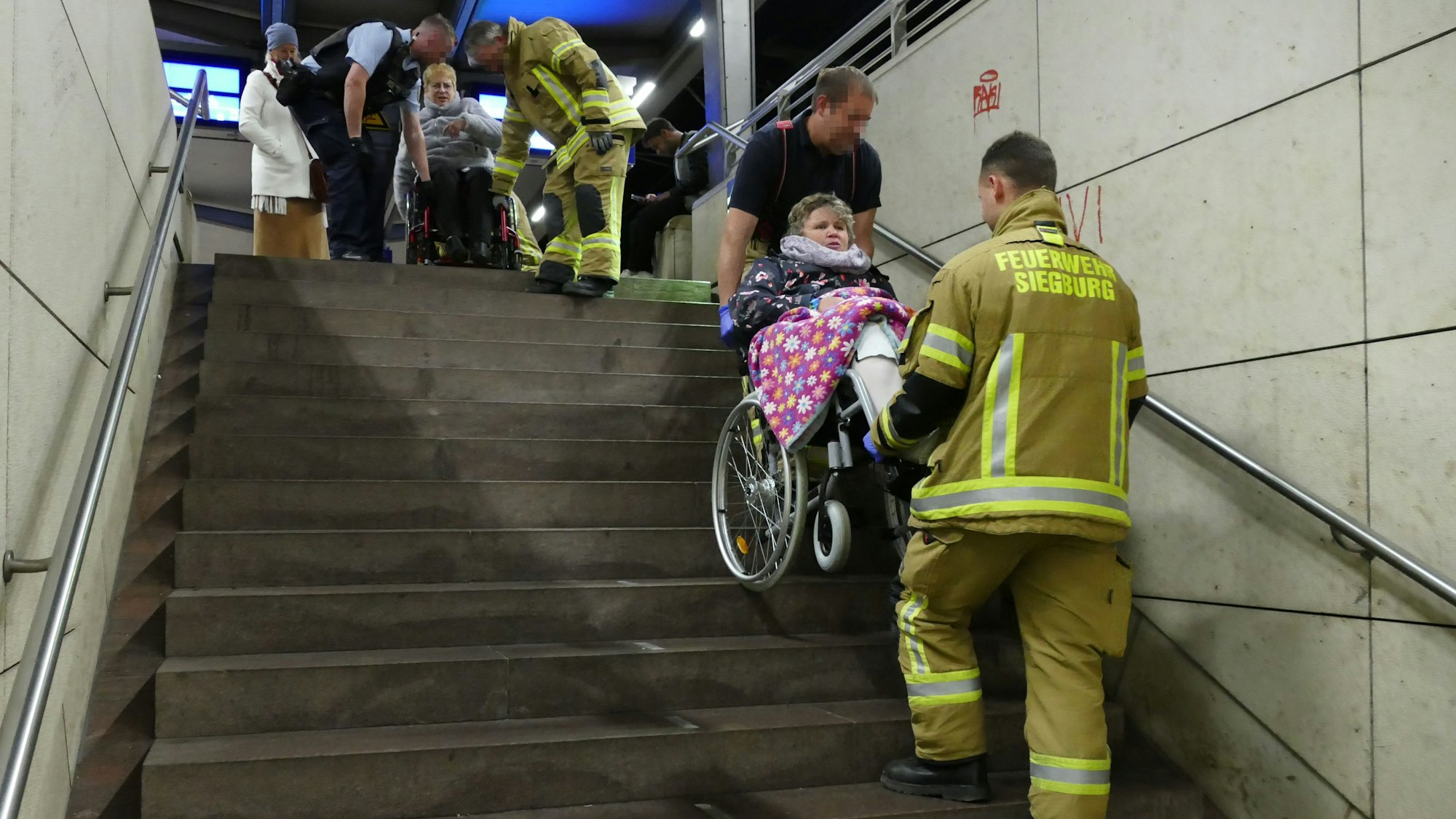 Zwei Männer in gelben Feuerwehranzügen tragen zwei Frauen im Rollstuhl die Treppe herunter.