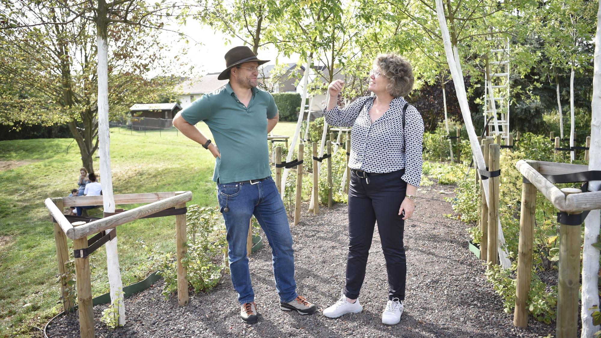 Ein Mann und eine Frau, Christoph Zimmermann und Anne Horst, stehen unter einer Reihe junger Bäume und unterhalten sich.