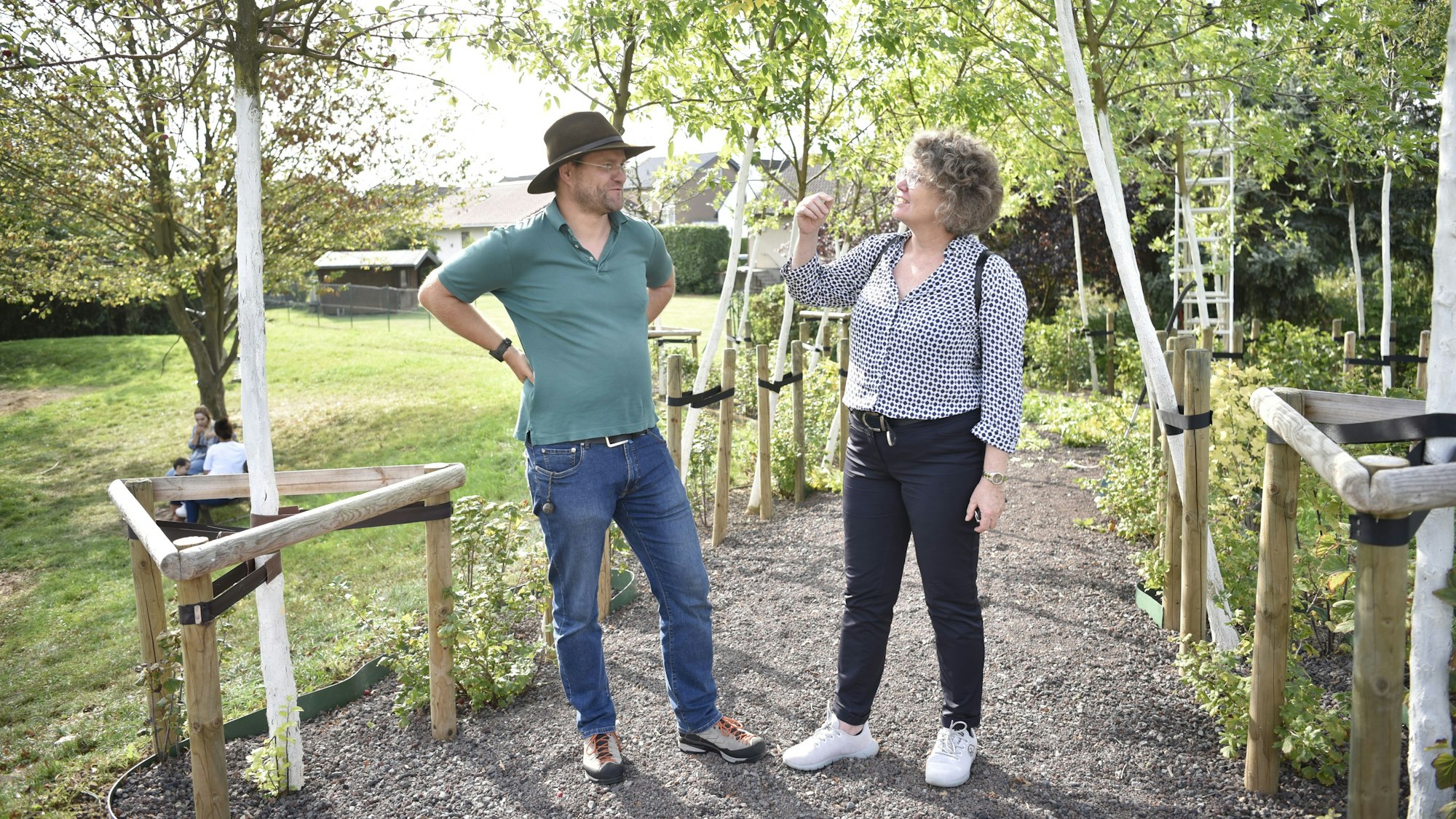 Ein Mann und eine Frau, Christoph Zimmermann und Anne Horst, stehen unter einer Reihe junger Bäume und unterhalten sich.