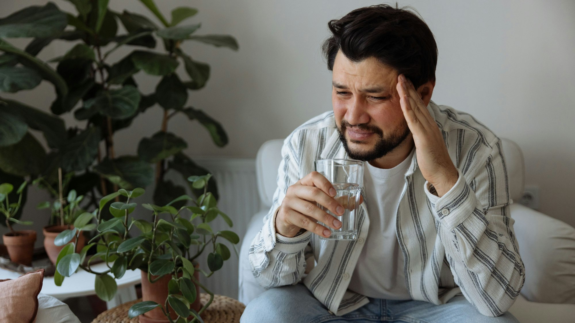 Ein Mann sitzt mit verzerrtem Gesicht im Wohnzimmer, hält mit der einen Hand seinen Kopf und in der anderen hand ein Glas.