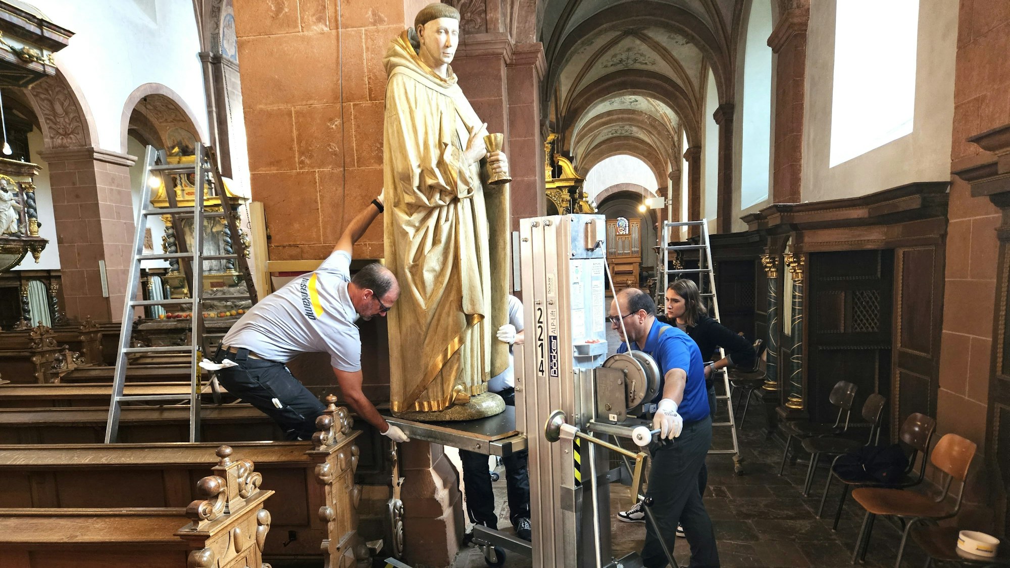 Die Figur des heiligen Hermann Josef steht auf einem Hubwagen, der von zwei Arbeitern bedient wird.